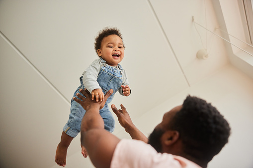 Loving father tossing his adorable laughing baby boy up in the air while playing together in their living room at home