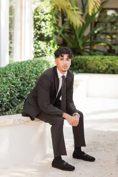 Photo of 2023 High-School Senior Young Man Well-Dressed in a Black Suit, White Dress Shirt, Black Tie & Black Suede Shoes, An18-Year-Old Cuban-American with Brown Hair & Brown Eyes Looking Fresh & Sharp in a Suit & Tie in a Classy Location in Palm Beach, Florida