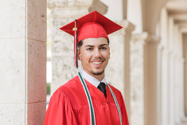 ein junger mann der high school 2023 in einer roten abschlussmütze und einem roten kleid mit drei ehrenquasten, ein gut gekleideter 18-jähriger kubanischer amerikaner mit braunen haaren und braunen augen, der in anzug und krawatte an einem noblen ort in  - men graduation clothing brown hair stock-fotos und bilder