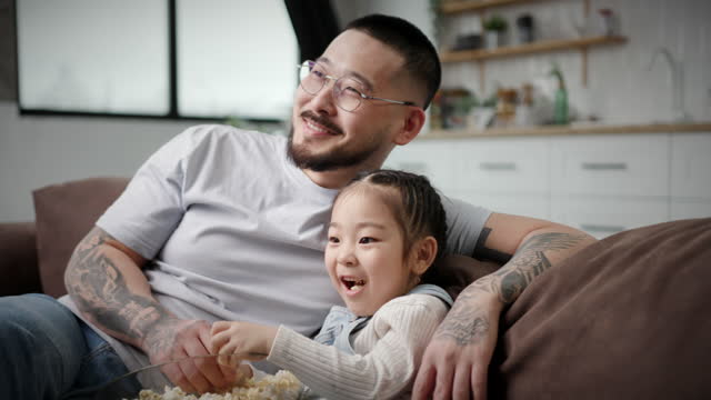Dad And Daughter Watching TV Eating Popcorn. Korean family having fun sitting on couch at home. Loving father hugging little daughter.