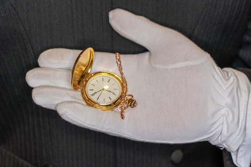 A hand in a white glove holding a gold pocket watch on a black background, close-up
