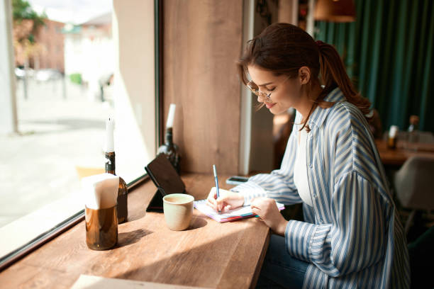 istock Portrait with concentrated young girl, student writing in notebook while learning with tablet and coffee cup in cafe. Freelance work, business people 1490980490