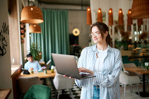 Smiling young woman, girl wearing eyeglasses, freelancer holding laptop and working with happy face in cafe. Social networks, freelance work, business people, remote job, career growth, ad concept