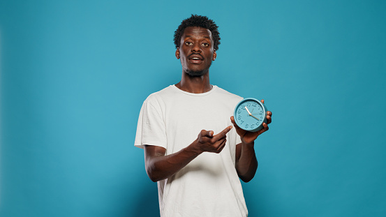Casual person pointing at time on wall clock for hour and minutes. Portrait of young man looking at watch and explaining countdown and delay for work. Punctual adult setting alarm