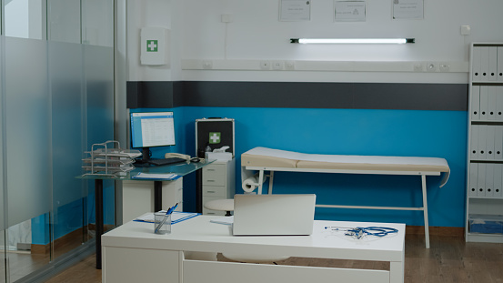 Nobody in doctors office with modern funiture at medical facility. Empty examination cabinet for healthcare specialist. Close up of white desk with laptop, stethoscope and pens