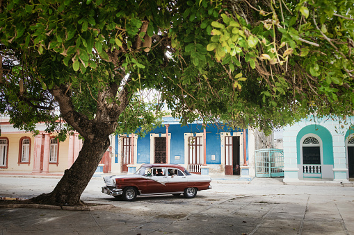 Parked in Gibara, situated in Holguin Province, an unrecognizable driver has his head turned while looking at the small town typical houses. In the meantime a senior woman, seated in the back seat of the vintage car, i.s looking the other way.