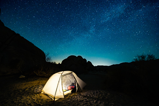This is a photograph of an illuminated tent at night with a Milky Way sky at the Indian Cove Campground in Joshua Tree National Park, located in the Mojave Desert in California, USA.