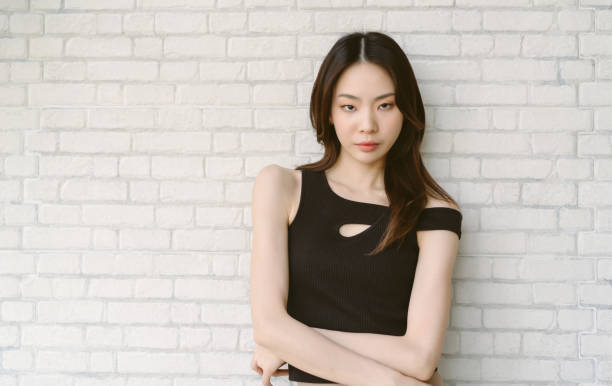 calm serious beautiful asian woman wearing off shoulder black and posing with crossed arms and looking at camera on white brick wall background. attractive young adult gen y lady face without smile. - korean culture fotos imagens e fotografias de stock