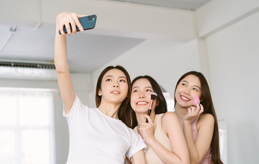 Portrait of three young Asian women with pretty, charming, attractive, cute, stylish, cheerful make-up in living room and making selfie on smartphone. Frenship and wellbeing concept