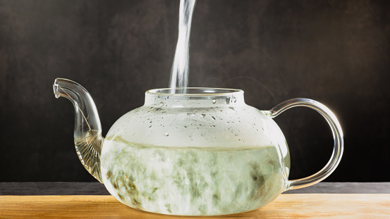 A beautiful glass teapot for brewing tea stands on a wooden board on a dark background, the teapot is transparent. Hot water is poured into the kettle, green tea is brewed, the effect of long exposure