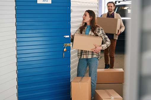 Lady is holding a large cardboard box and opening the door of the pantry in the storage warehouse. In the background, a guy carrying a box with things from car