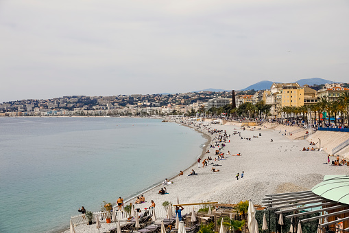 Nice, France - April 22, 2023: A view along the seashore of the city, its buildings and the beach where there are people despite the cloudy day.