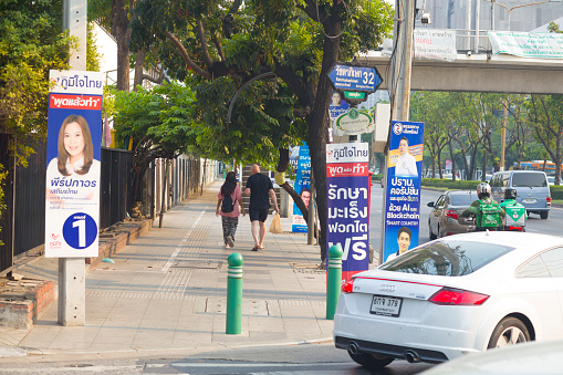 Thai woman and white bald man are walking on sidewalk of Ratchadaphisek Road in Bangkok Chatuchak. On street is some traffic. On sidewalk are banners with candidates for election. In background is pedestrian footbridge