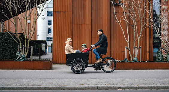 Portrait of a little girl sitting in the front cart with her mother while her father riding a cargo bike on the city street.