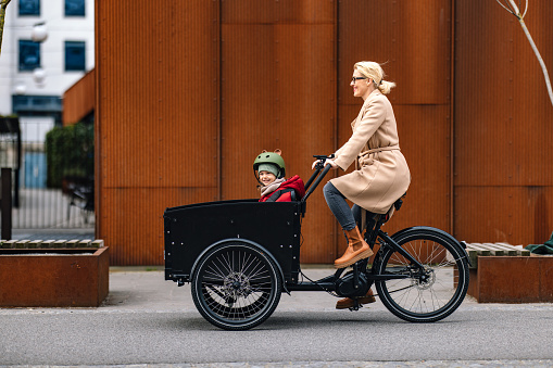 Side view of a smiling woman having fun riding a cargo bike on the city street with her beautiful daughter sitting in the front cart.