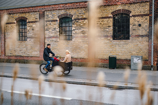 A little girl sitting in the front cart with her mother while her father riding a cargo bike on the city street.