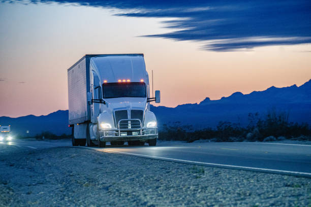 a white semi truck traversed the nevada desert, its silhouette cutting through the vast expanse. against the backdrop of the setting sun, the majestic mountains rose, casting shadows in shades of purple and orange. - truck desert semi truck orange imagens e fotografias de stock