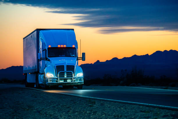 a white semi truck traversed the nevada desert, its wheels kicking up dust as it journeyed through the vast expanse. against the backdrop of majestic mountains, the setting sun cast a warm glow, painting the scene with hues of orange and purple. - truck desert semi truck orange imagens e fotografias de stock