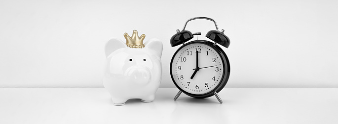 Black alarm clock with piggy bank against a white wall. Financial concept.