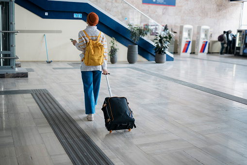 Rear view of a female tourist with luggage woman walking on railway station hallway. Woman going on vacation walking on train station with bags.