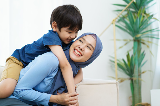 Happy cheerful Asian muslim family living together in a weekend, woman muslim embracing her lovely little son and smiling to camera. Modern muslim lifestyles concept.