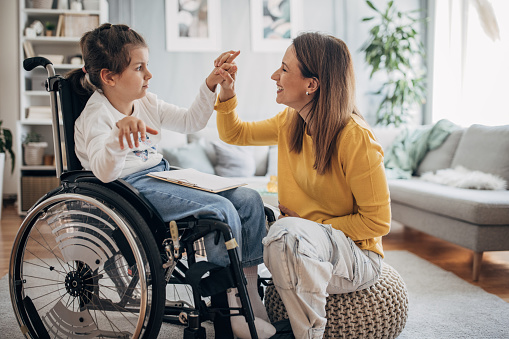 Mother and young daughter who is in wheelchair enjoying together in living room at home.