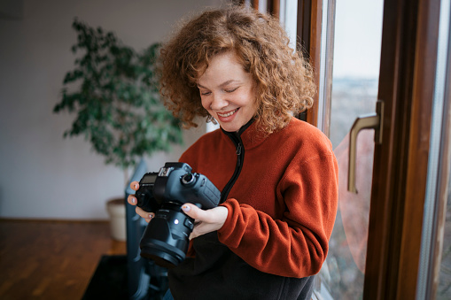 Young woman looking photos on her dslr camera and smiling. Female photographer standing by window and holding dslr camera at home office.