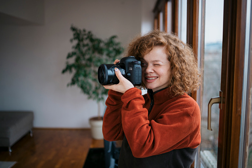 Woman photographer working in home studio testing her new dslr camera. Young female taking photograph with her professional dslr camera standing by window at home office.