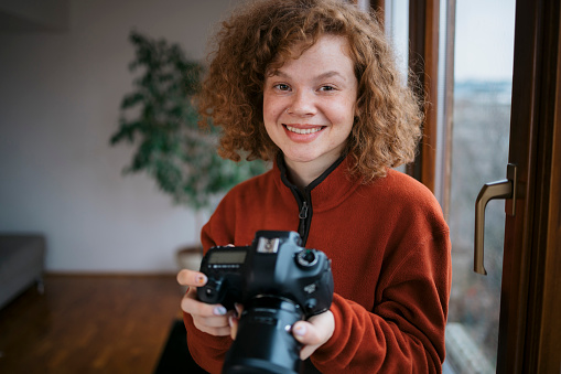 Portrait of a happy young woman with dslr camera standing by window at her home studio. Creative female photographer holding her professional digital camera.