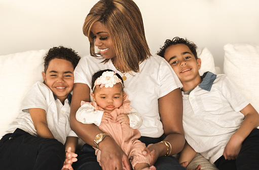 View of beautiful African American woman  sitting on couch with her smiling six year old twin boys and baby daughter on her lap; light wall in background