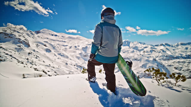 SLOW MOTION Full length of man with snowboard admiring landscape. Male snowboarder is wearing warm clothing. He is standing at peak on sunny day.