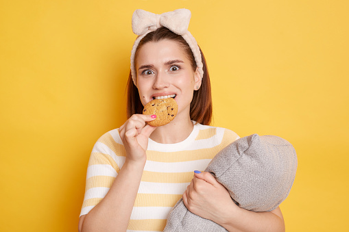 Confused woman wearing striped T-shirt and hair band standing isolated over yellow background posing with pillow in hands biting delicious cookie looking at camera.