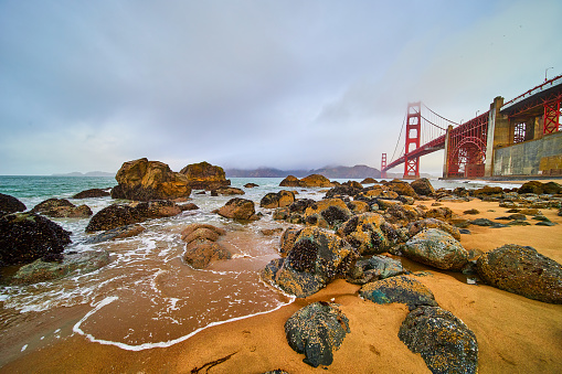 Image of Stunning sandy beach waves crashing into rocks covered in mussels by Golden Gate Bridge on foggy morning