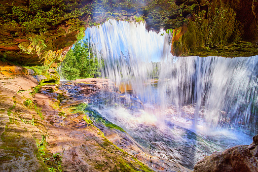 Image of View behind and inside of waterfall cave of orange and lichen growth with sun hitting the water