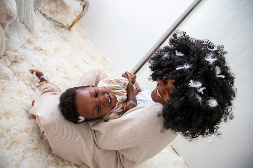 Beautiful young African American mom playing with her black little son. They smiling and having fun in soft white feather in the kid's room floor. Free copy space