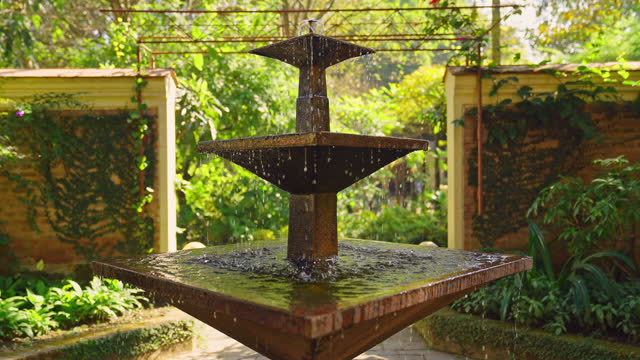 Moving towards an outdoor water fountain feature in slow motion with garden greenery and brick wall in the back