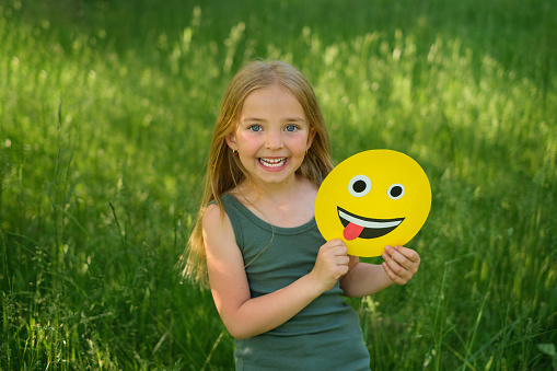 A long-haired blonde girl laughing merrily holds a paper emoticon face in her hand, showing her tongue. Celebrating the World Smile and Emoji Day