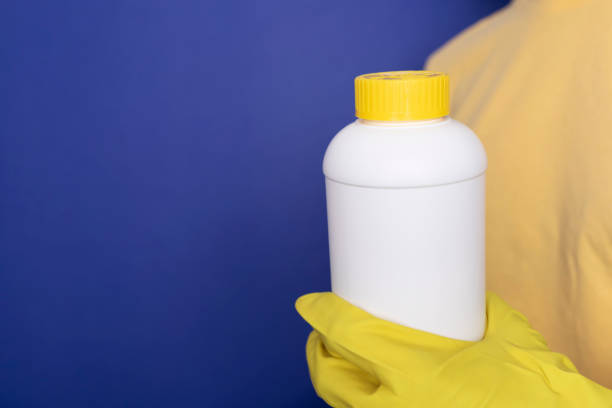 Woman Hand In Yellow Gloves Holds White Bottle With Pipe Cleaner Granules. Removal Of Blockage In Sink, Shower With Special Chemicals. Cleaning Clog In Kitchen, Bathroom. Blue On Background,Copyspace la mano de la mujer en guantes amarillos sostiene una botella blanca con gránulos de limpiador de tuberías. eliminación de obstrucción en el fregadero, ducha con productos químicos especiales. limpieza de obstrucciones en cocina, baño. azul en el fon