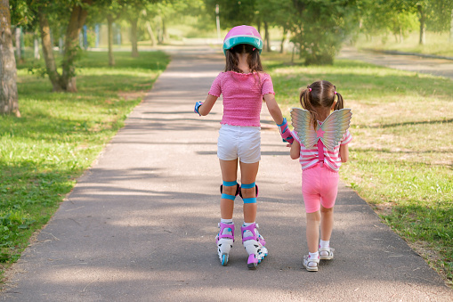 A little girl leads her older sister, who is learning to rollerblade, by holding her hand.  A child in a suit with wings over his shoulders