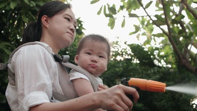 A mother holds her 7-month-old baby using a water nozzle for watering the plants