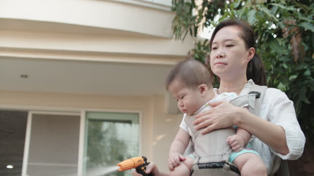A mother holds her 7-month-old baby using a water nozzle for watering the plants