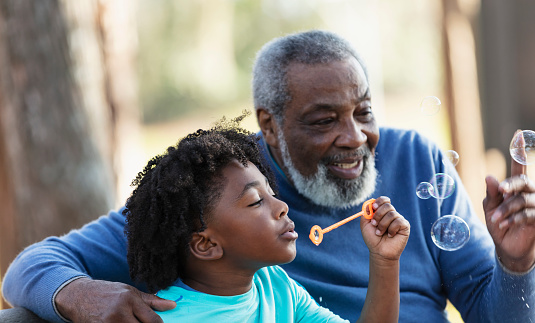 Headshot of an African-American boy and his grandfather having fun together at the park, sitting on a park bench, blowing bubbles with a bubble wand. Grandpa is smiling and trying to pop a bubble, with his arm around his grandson's shoulders. The main focus is on the boy, who is 5 years old.