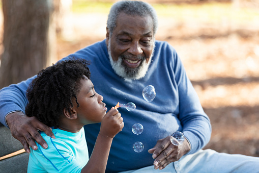 An African-American boy and his grandfather having fun together at the park, sitting on a park bench, blowing bubbles with a bubble wand. Grandpa is smiling and watching, with his arm around his grandson's shoulders. The main focus is on the boy, who is 5 years old.
