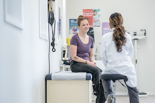 A young woman sits up on an exam table during a medical appointment.  She is dressed casually as she talks with her doctor about her health concerns.