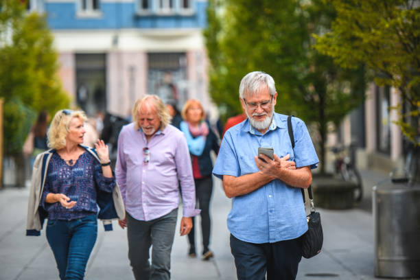 Senior caucasian male texting with a smart phone while walking in the city. Defocused senior adults walking in the background.