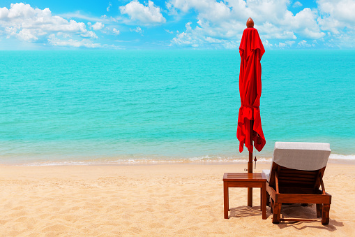 Red closed umbrella, empty deck chair, table, folding solar parasol, sun lounger, wooden chaise lounge, sun bed, paradise tropical island sea beach background, summer holidays, relax vacation, travel