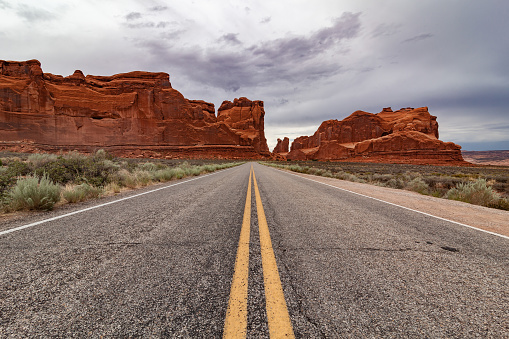 Highway to the world of arches. The red mountains in the background, bushes on the roadside, to go to an enchanted world. Arches National Park is a national park in eastern Utah in the United States. The park is adjacent to the Colorado River 4 miles (6 km) north of Moab, Utah.