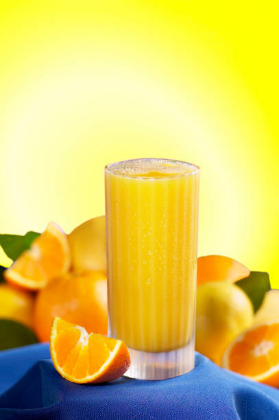 un vaso estriado lleno de jugo de naranja con gotas de agua y condensación descansando sobre un mantel azul con una cuña naranja y una variedad de cítricos sobre un fondo azul. - orange portion fruit citrus fruit fotografías e imágenes de stock