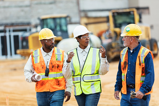 A African-American woman in her 30s working at a construction site, wearing a hardhat, safety goggles and reflective vest. She is looking over the camera with a confident expression, smiling, showing thumb up.