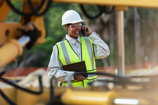 A mature African-American woman, in her 40s, wearing a hard hat, safety vest and safety goggles, working at a construction site. She could be a construction worker, engineer, or building contractor. She is holding a digital tablet, talking on her mobile phone.